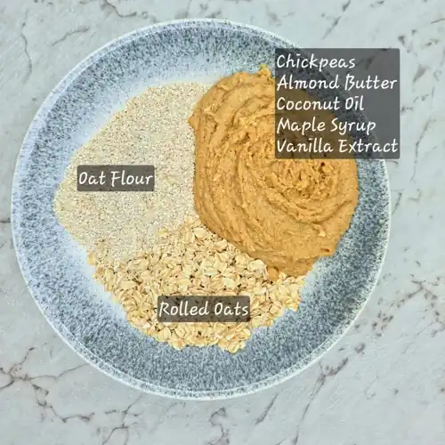  Add rolled oats and oat flour to chickpea puree mixture. Chickpeas, almond butter, coconut oil, maple syrup and vanilla extract. Labelled recipe ingredients. 